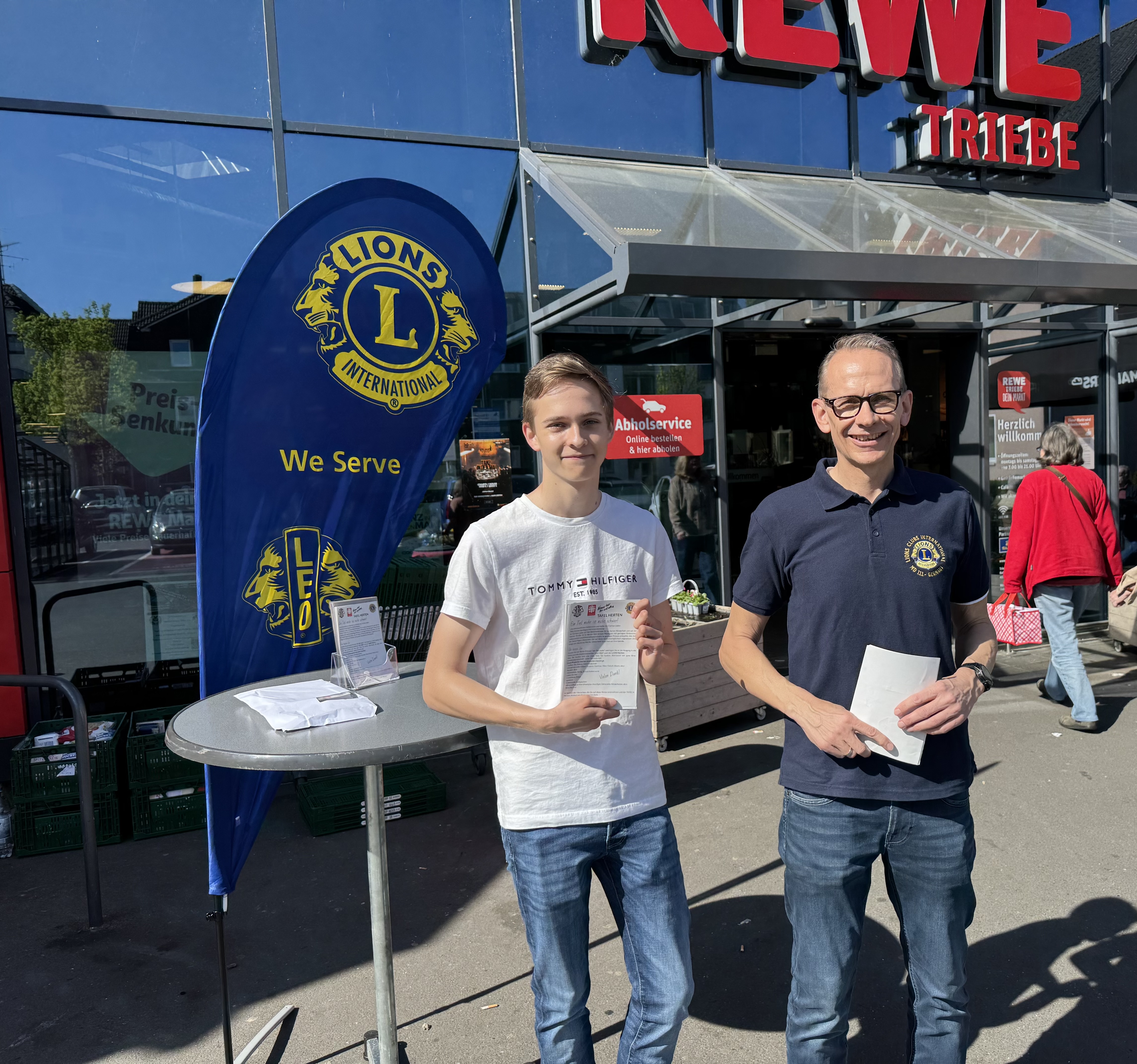 Das Bild zeigt ein Mitglied der LEOS und ein Mitglied der  LION vor dem REWE Markt Triebe in Herten Westerholt für die Ein teil Mehr Aktion im April 2025